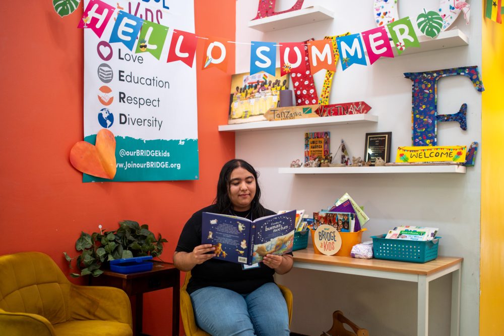 Joselyn Moreno reads a book underneath a "Hello Summer" banner at ourBRIDGE for KIDS