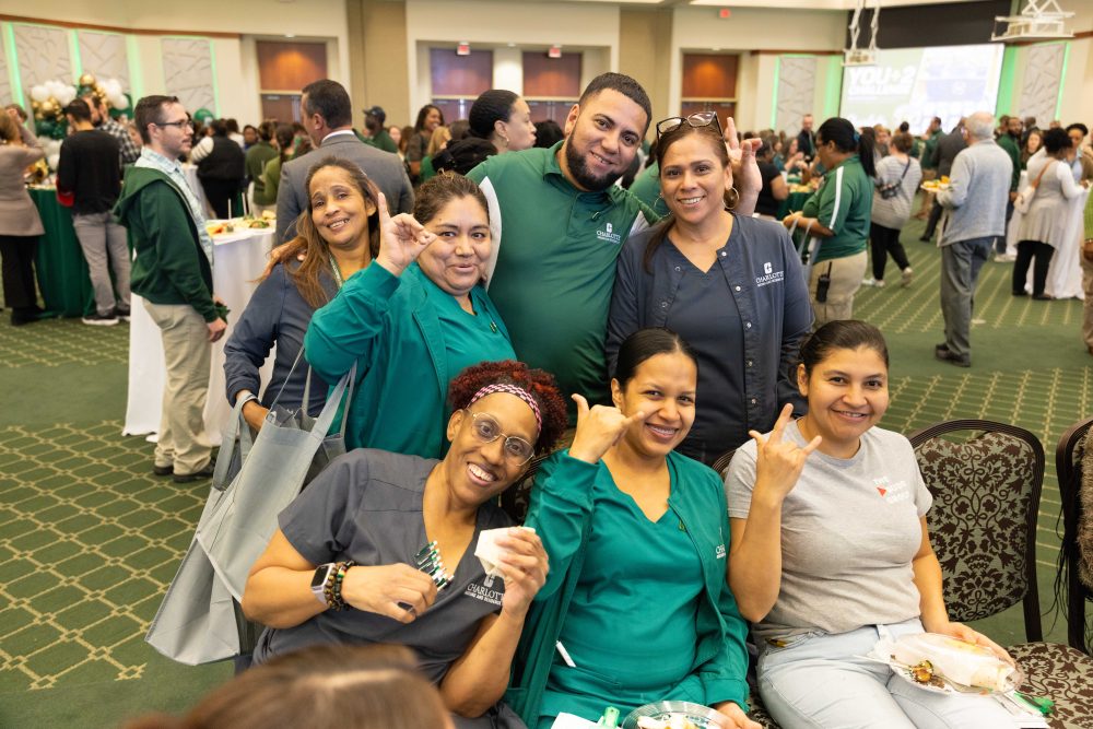 A group of smiling healthcare workers in green scrubs and casual attire pose together at a festive event, making playful hand gestures while seated in a large hall with other attendees and food tables in the background.