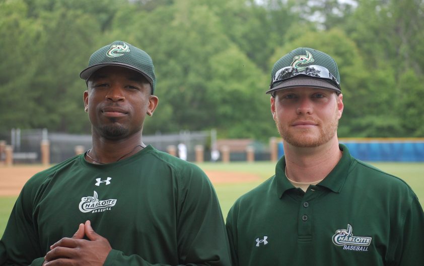 Two Charlotte baseball coaches stand side by side on the baseball field wearing green Charlotte Baseball shirts and matching team caps, posing for a portrait with the field and trees in the background.