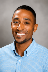 2026 Truist Report - Banks Headshot Professional headshot of a smiling man with short dark hair and a trimmed beard, wearing a light blue button-down shirt against a softly blurred gray background.