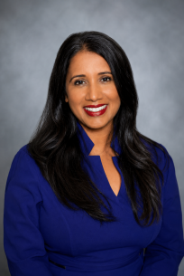 2026 Truist Report - Gooty Headshot Professional headshot of a smiling woman with long dark hair, wearing a royal blue dress and red lipstick, posed against a gray studio backdrop.