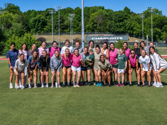 2026 Truist Report - SELP A large group of athletes pose together on a campus soccer field after a training session. Everyone smiles for a group photo with the Charlotte athletic facilities and tree-lined campus visible in the background.