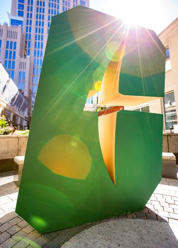 Large green “C” sculpture in an urban plaza with sunlight streaming through its center, creating lens flare, with tall buildings in the background.