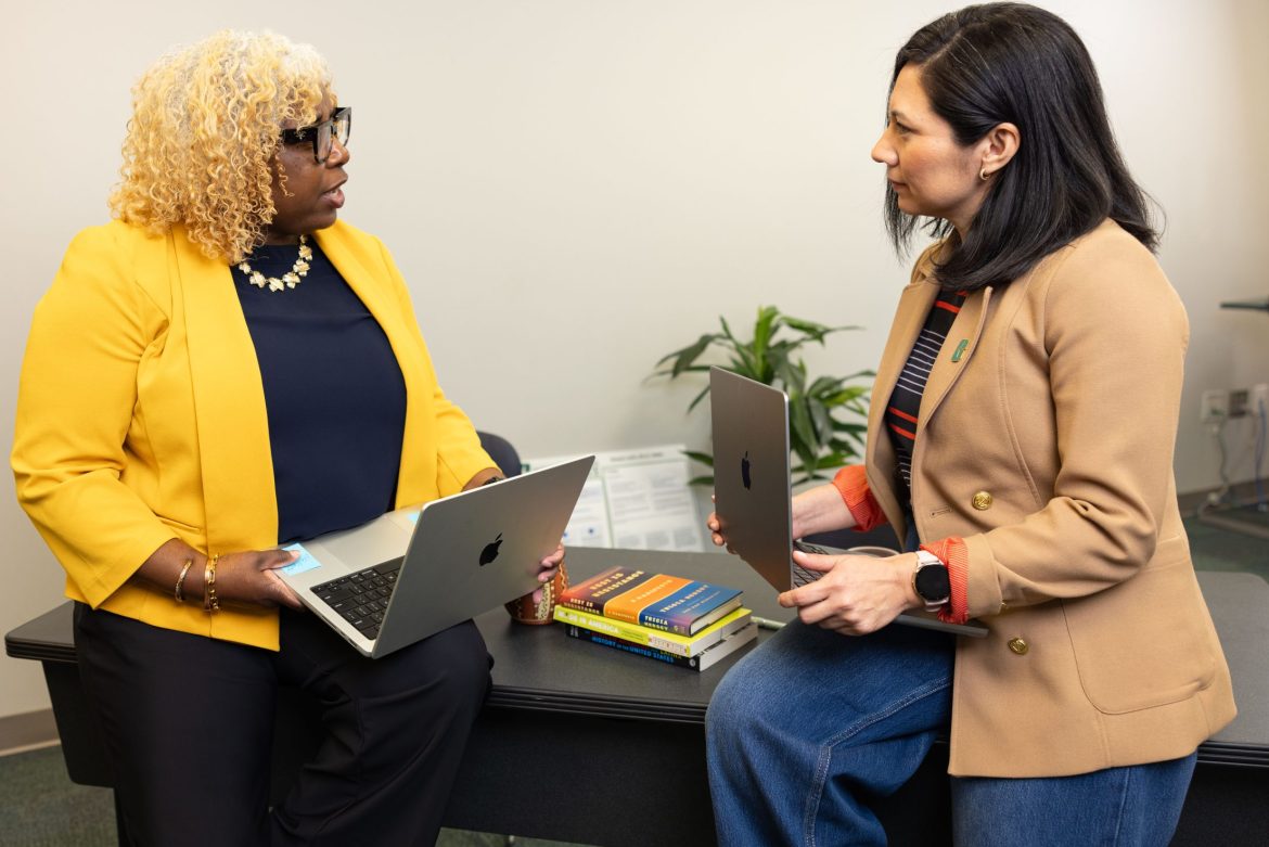 Two women holding laptops sit and talk in an office setting beside a desk with stacked books and a small plant.