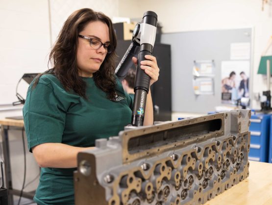 Engineer operates a handheld device to measure an engine part on a wooden table in a lab setting.