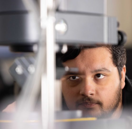 Man closely examines machinery, focusing on equipment during a lab experiment with blurred foreground elements.