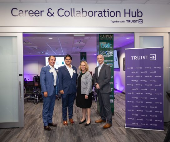 CareerCenter+Truist_23105_2E7A2849_ah Four professionally dressed individuals pose at the entrance of the Career & Collaboration Hub, sponsored by Truist. A Truist banner stands beside the group, with meeting rooms and employer partner signage visible inside the career center.
