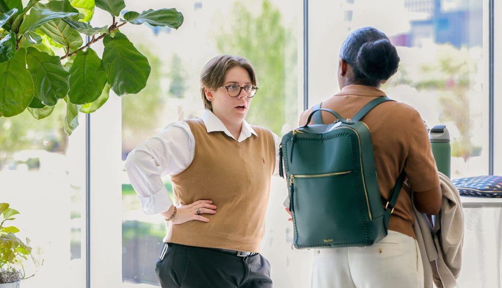 Two women talking indoors near large windows, one gesturing while speaking as the other listens with a backpack.