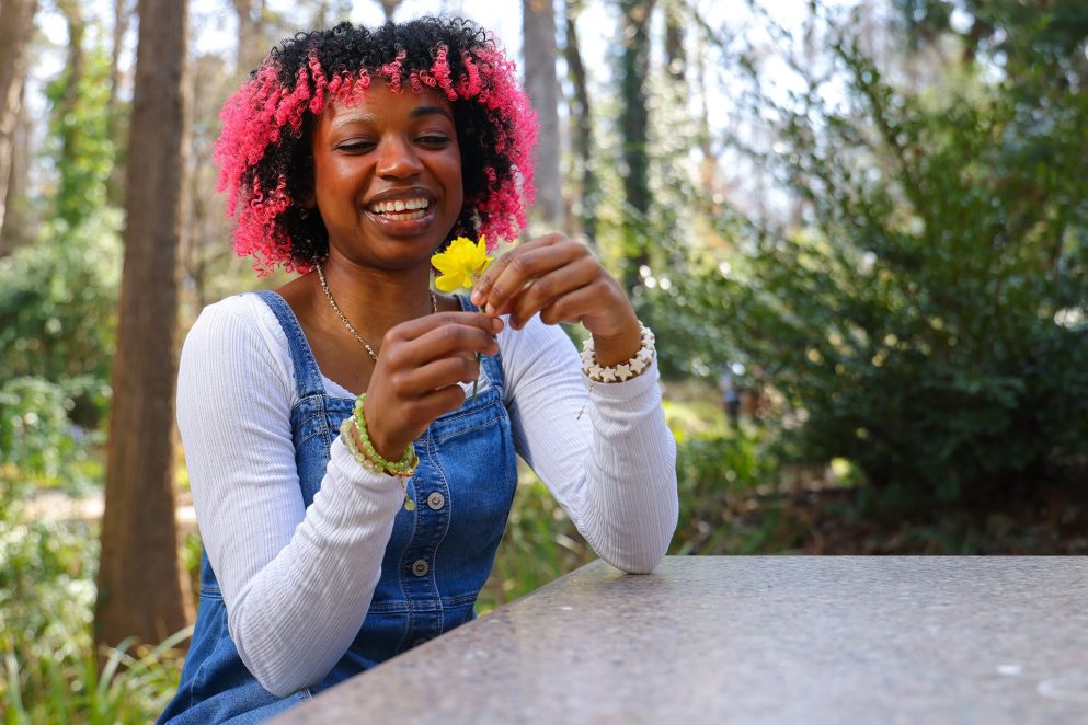 Student with curly pink-highlighted hair smiling while holding a yellow flower at an outdoor table surrounded by trees.