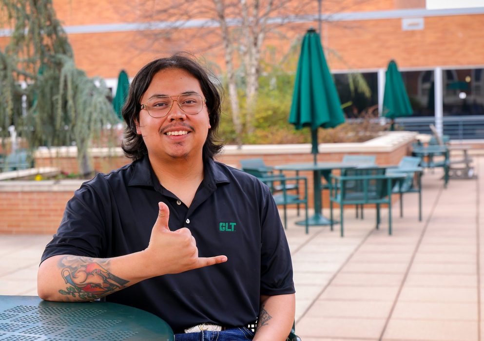 Transfer student sits at an outdoor campus table, smiling and making a hand gesture, with patio seating and campus buildings in the background.