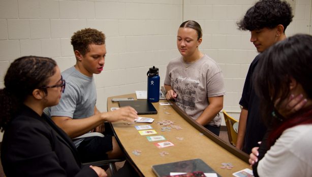 A group of college students gathered around a curved classroom desk, playing the board game PLACES. Game cards and tokens are spread out on the table, and a laptop and water bottle are also visible. The classroom has white brick walls and a clock on the wall. {{brizy_dc_image_alt imageSrc=