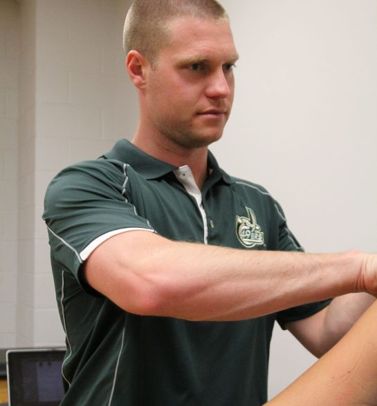 An athletic trainer wearing a green 49ers polo evaluates a student's shoulder and arm during a clinical assessment in a sports medicine laboratory.