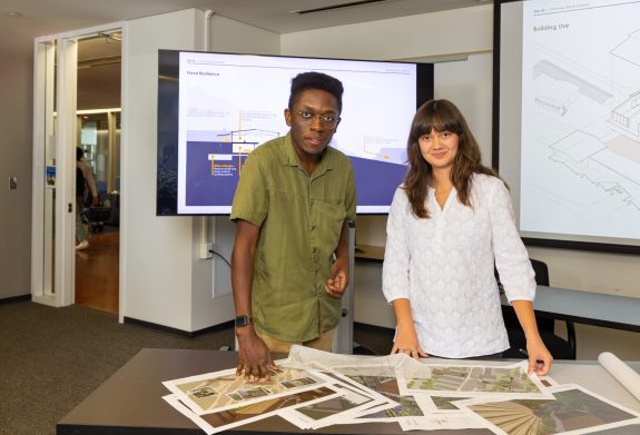 Two students stand at a table with printed design plans, presenting their project in front of screens displaying diagrams and renderings.