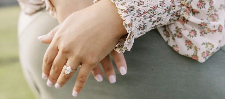 Close-up of a person's hand with a diamond ring resting on the back of someone wearing a green shirt, set against a blurred outdoor background.