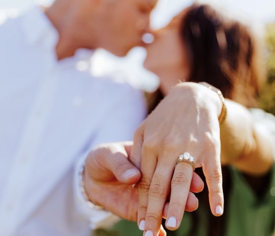 Close-up of a woman’s hand with an engagement ring as a couple kisses in the background.