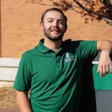 UNC Charlotte student in a green campus polo poses casually outdoors by a campus sign. {{brizy_dc_image_alt imageSrc=