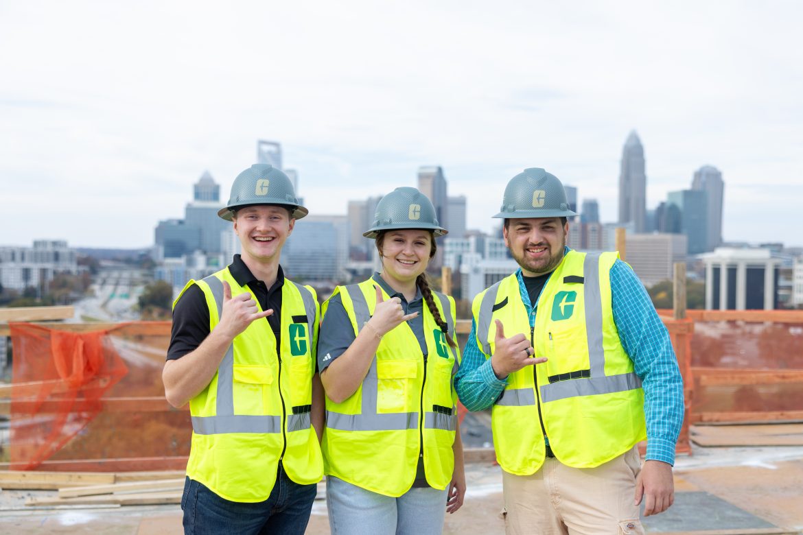 Renegade-BTS-9968 Three construction students stand in hard hats in front of the Charlotte skyline