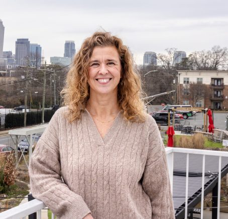 Woman smiling on a balcony with the Charlotte skyline in the background on an overcast day.