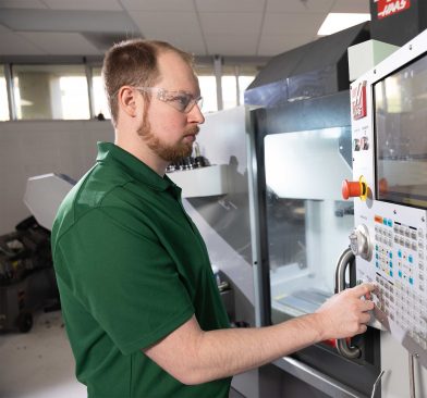 Person wearing safety glasses operates a CNC machine, adjusting controls on the interface panel in a lab or workshop setting.