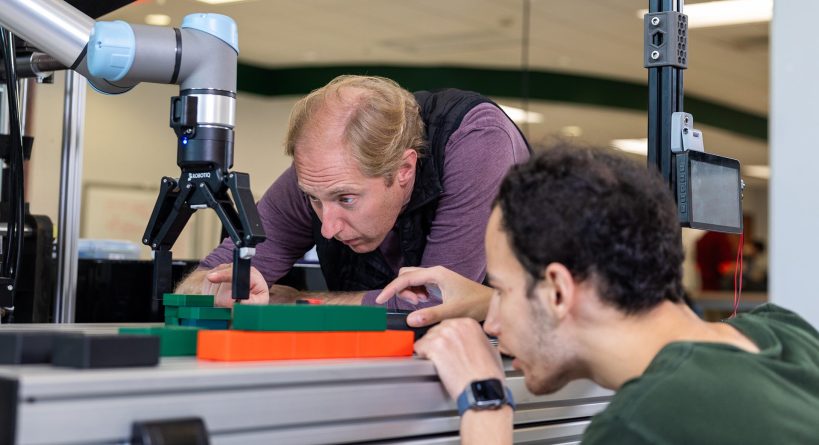 Two people closely inspect and adjust a robotic arm working on a small assembly in a lab setting.