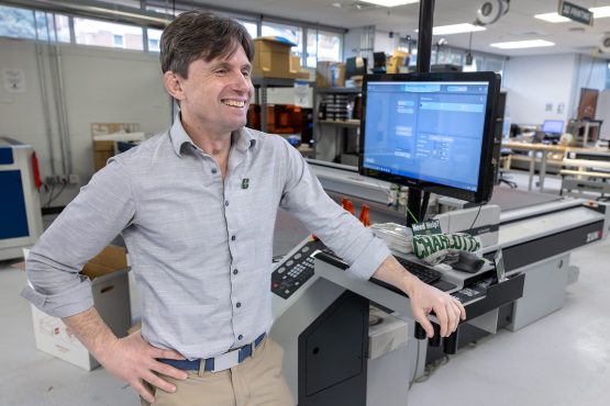 Person stands smiling beside a digital fabrication machine and computer workstation in a university lab filled with advanced equipment.