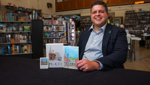 A smiling man in a UNC Charlotte lapel pin sits at a table in a board game store, showcasing a card game titled {{brizy_dc_image_alt imageSrc=