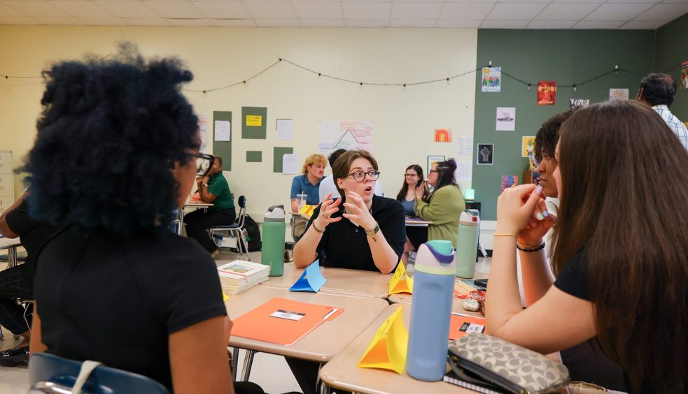 Instructor leading a discussion with students seated in small groups around desks in a classroom.