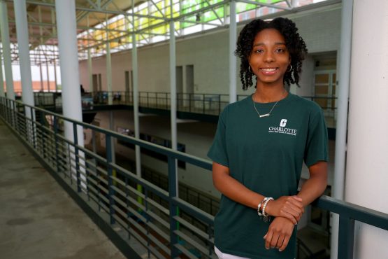 Smiling student in a green UNC Charlotte T-shirt stands in a modern campus atrium, leaning on a railing with open walkways and structural beams in the background.