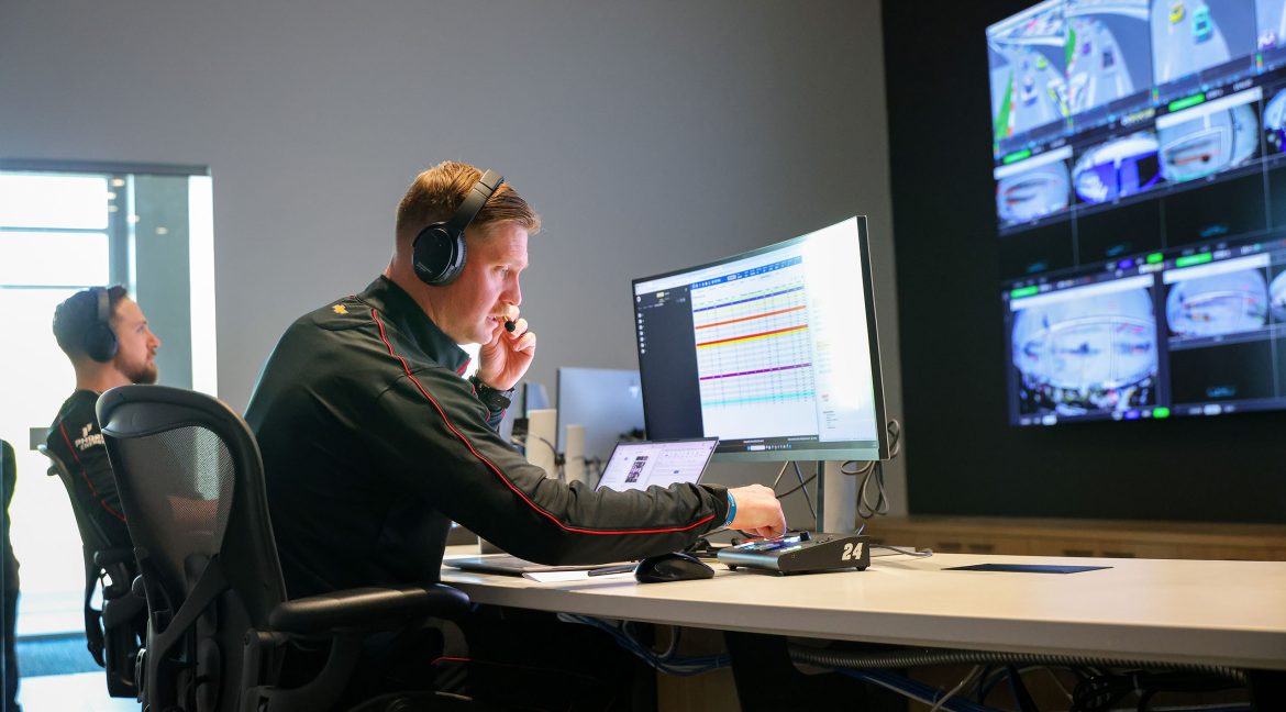 A Hendrick Motorsports engineer uses a headset and multiple monitors in a race operations center while reviewing live timing, strategy data, and track camera feeds.