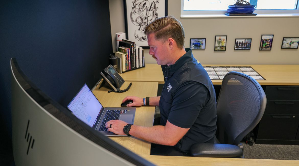 A Hendrick Motorsports engineer works at a laptop in a quiet office, analyzing performance data at a desk with books, framed photos, and a telephone.