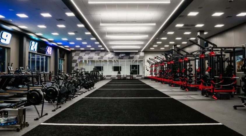 Wide view of the Hendrick Motorsports performance center featuring indoor sprint lanes, weight racks, rowing machines, and cardio equipment in a spacious gym.