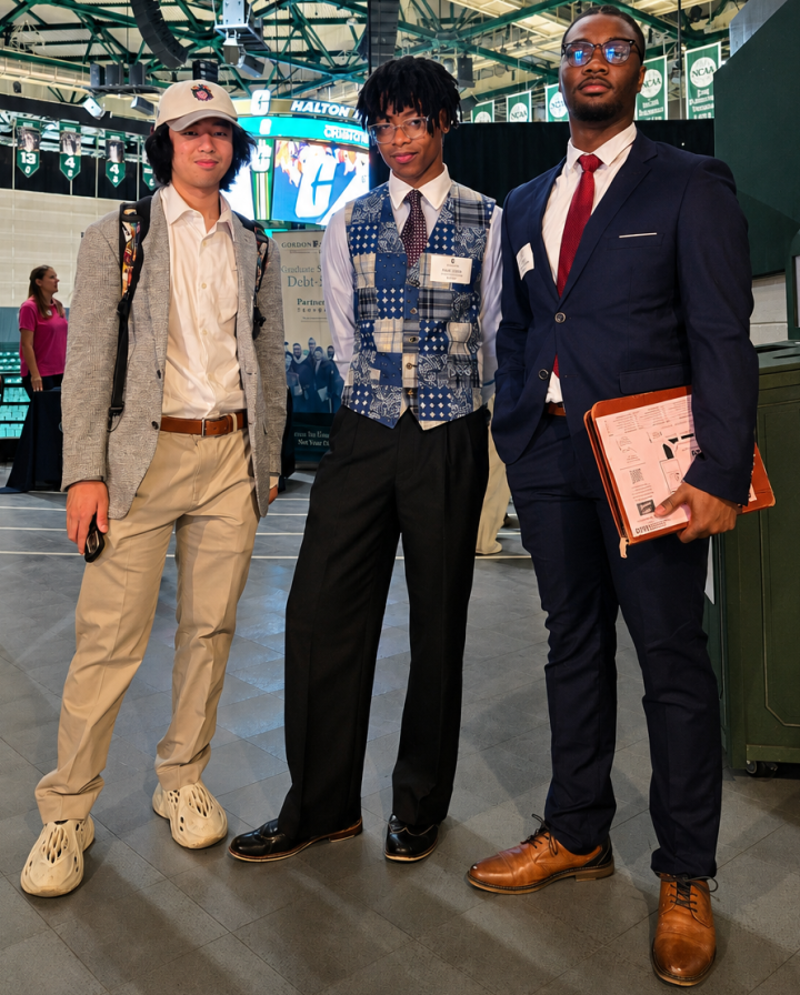 Truist Image 2 Three professionally dressed students pose together at a career fair in a university arena, standing beneath championship banners and holding resumes and networking materials.