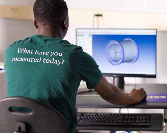 Student at a computer works on a 3D model, wearing a shirt that reads, “What have you measured today?”